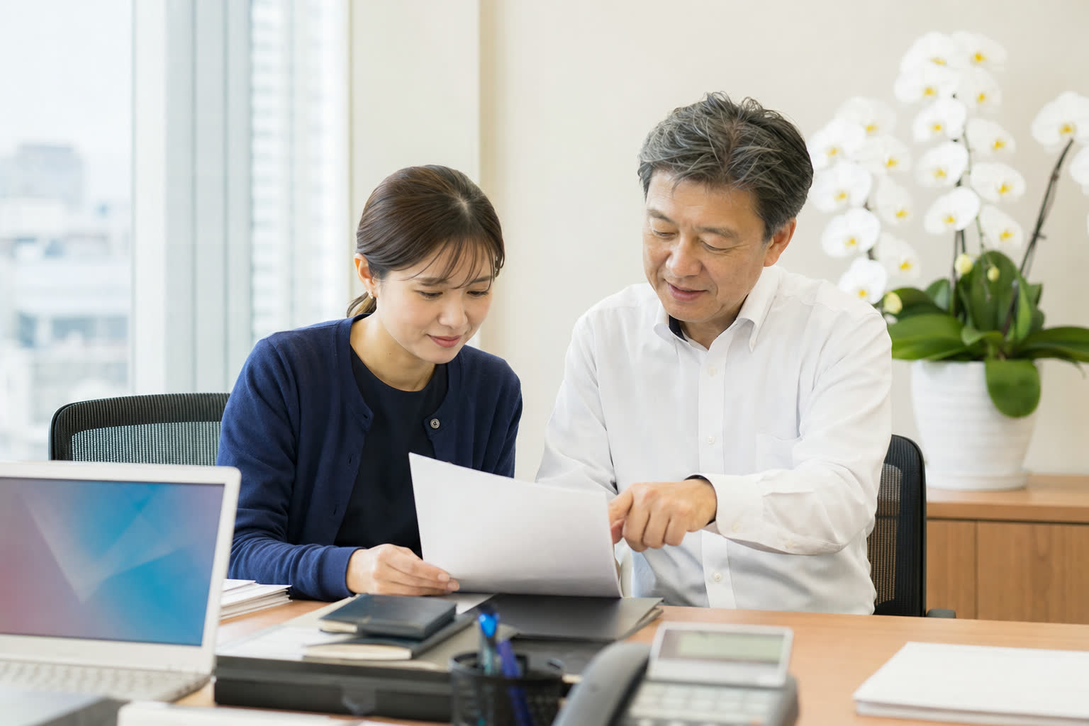 オフィスの机で上司と担当者が書類を確認し、奥に白い胡蝶蘭が置かれている