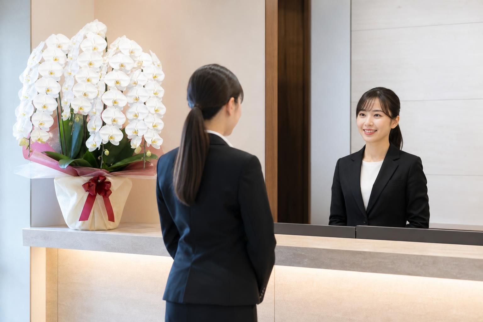 会社の受付に飾られた白い胡蝶蘭の5本立と、受付の社員にあいさつする来社した女性